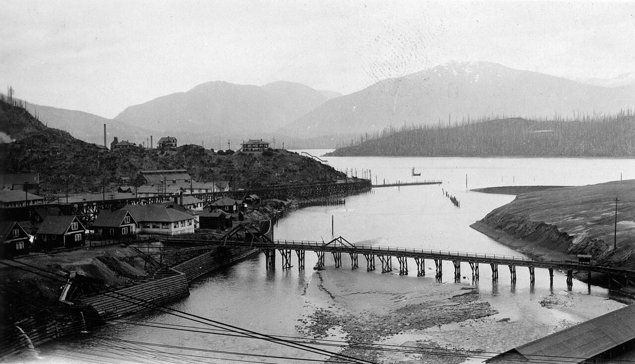 Black-and-white photo of a long wooden bridge across an inlet, with buildings and mountains
