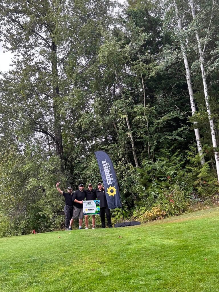 A team of golfers poses with a sign beside a sponsor banner at the edge of the trees.
