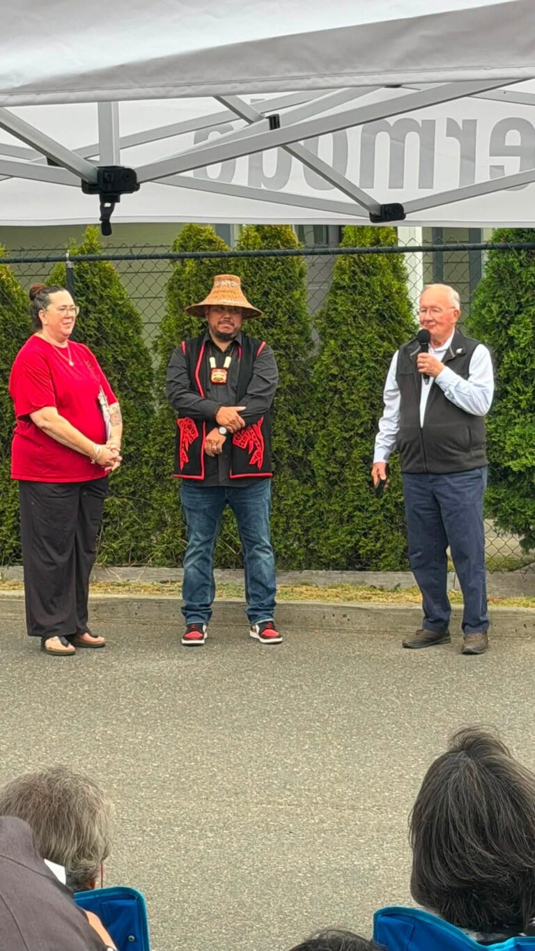 Three people stand in front of the audience at the celebration; one speaks into a microphone.