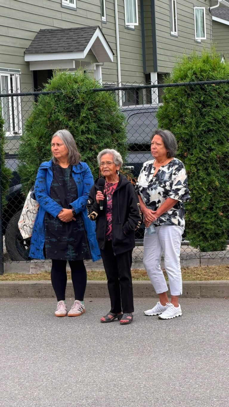 Three women stand together in front of a hedge at the celebration.