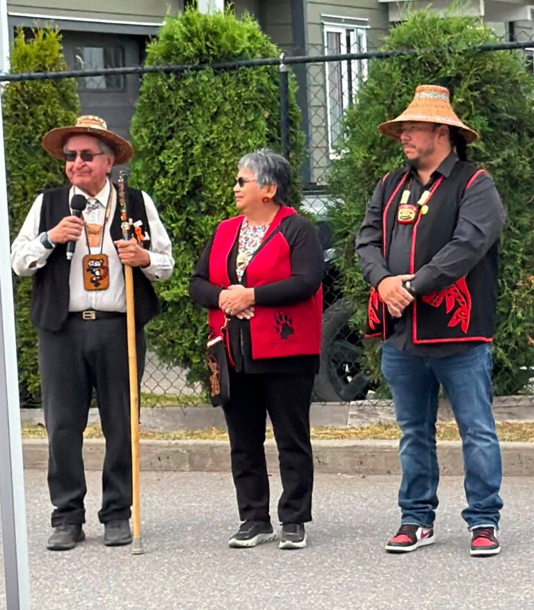 Three people in regalia stand in front of a hedge; one speaks into a microphone.