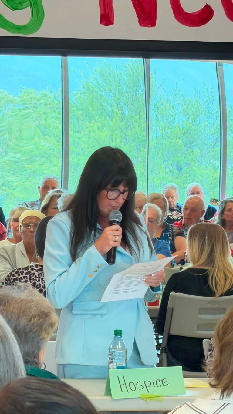 A woman in a light blue suit reads from notes at a microphone in front of a seated audience.