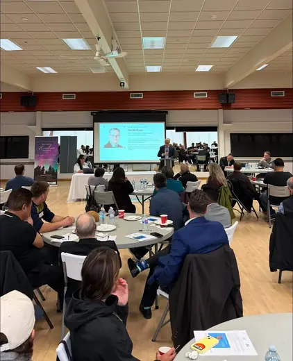 A full room of attendees at round tables facing the presentation screens.