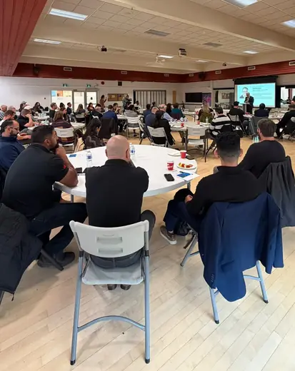 View from the back of the room: attendees at round tables and presentation screens at the front.