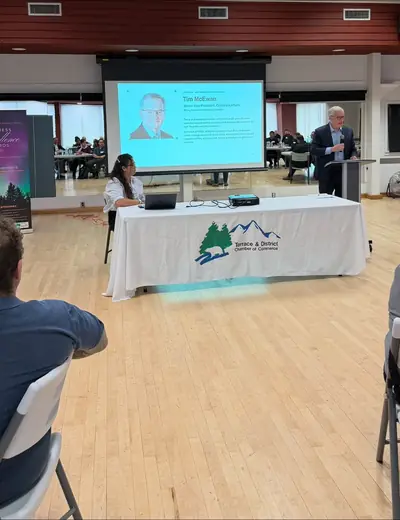 A speaker at a podium beside a table with a Terrace & District Chamber of Commerce banner; a speaker photo is on the screen.