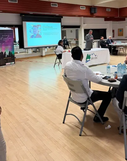 A speaker stands at the front of the room while attendees at round tables listen.