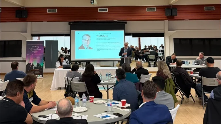 Attendees at round tables watch a speaker at a podium beside a large projection screen at the front of the Terrace Sportsplex hall.