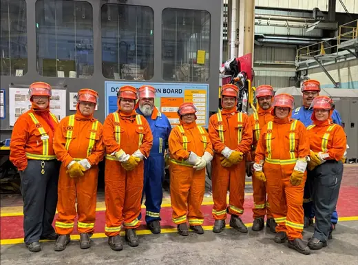 Tour group in orange and blue safety coveralls and hard hats standing in a row inside Rio Tinto BC Works.
