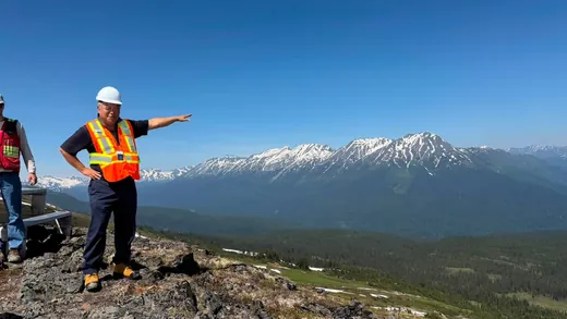A person in a white hard hat and orange high-visibility vest points out over a forested valley toward a range of snow-capped mountains at the KSM Project site.