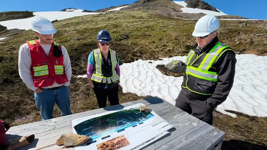Three people in hard hats and safety vests look over a printed project map laid on a wooden table on an alpine slope, with snowfields behind them.