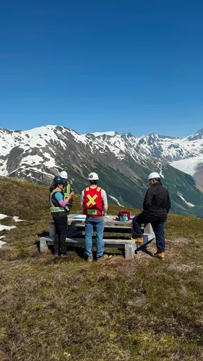 Four people in hard hats and safety vests stand around a picnic table on an alpine ridge under a clear blue sky, with snow-capped mountains and a glacier beyond.
