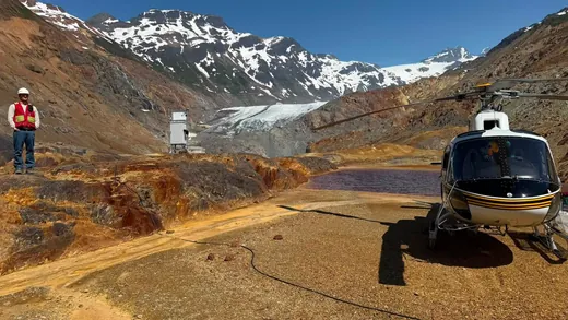 A helicopter parked on gravel beside a small pond in a rocky, orange-stained valley below a glacier and snow-capped peaks, with a person in a hard hat and red vest standing on a ridge at left.
