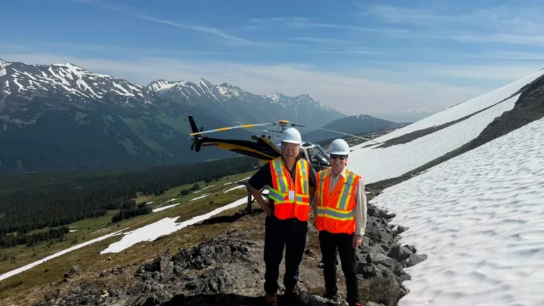 Two people in white hard hats and orange high-visibility vests stand on a rocky, snow-patched slope in front of a parked helicopter, with snow-capped mountains behind them at the KSM Project site.